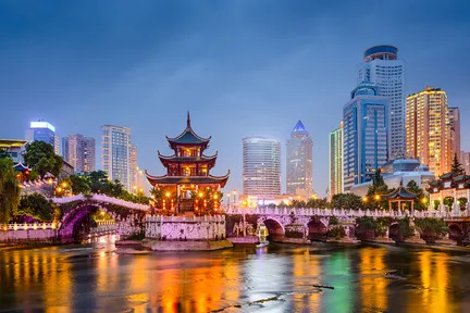 A dusk view of a traditional Chinese pavilion illuminated by warm lights, standing on a stone bridge over a river, with modern skyscrapers lit in the background.