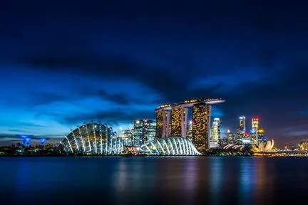 A nighttime view of Singapore’s waterfront with the illuminated Marina Bay Sands hotel, the curved glass domes of Gardens by the Bay, and skyscrapers reflecting in the water under a deep blue sky.